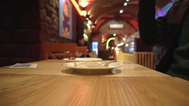 Closeup view of empty dessert plate with spoon and person placing empty tall glass on polished wooden table in atmospheric cafe. Warm lighting illuminates interior.