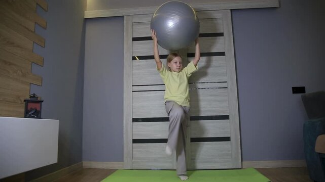 Caucasian girl performing indoor fitness routine, holding large gray stability ball above head, exercising on bright green mat, promoting strength and balance