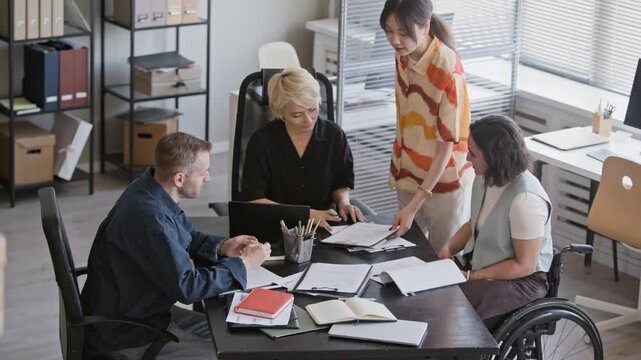 High angle stab shot of young Asian manager bringing document to middle-aged Caucasian employees and woman in wheelchair coworking in modern office