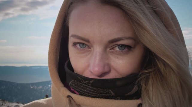 Portrait of a lovely girl enjoying ski season vacations. Blond hair moving in the wind in slow mo. Dressed in a hoodie, she fixes her hair with her hand. Mountain peaks in the snow. Winter travel.