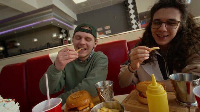 Medium high-angle shot of three joyous students toasting with fries, celebrating end of semester or school year at fast food restaurant, then tucking into tasty burgers with soda and milkshake