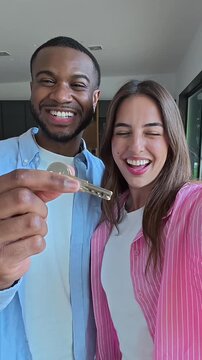 Happy young interracial couple smiling and celebrating while showing the keys to their new home, capturing a selfie video to share their excitement about moving in together and becoming homeowners
