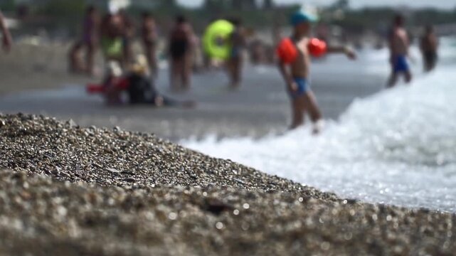 Blurred background showing many people on a vibrant beach, child wades in shallow ocean waves, families relax by sea, summer travel leisure, dynamic water motion, relaxing atmosphere, holiday scene. L