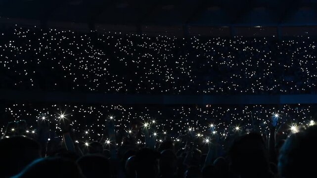 Excited crowd waving cellphones and flashlights at stadium during rock concert and sports game