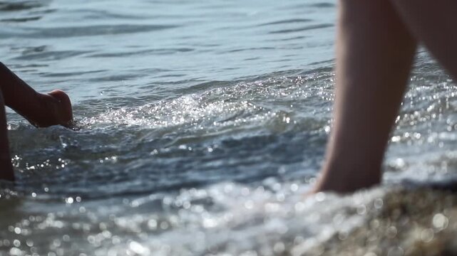 Close up view of person legs and feet walking slowly along pebbly shore line, gentle ocean waves caressing skin, shimmering sunlight creating bokeh effect on water surface. Vacation holiday leave in T