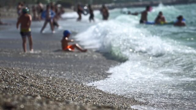 Waves washing over pebbly shore, tranquil. Background numerous blurred people enjoying ocean during sunny summer day. Children play in shallow water as adults swim and relax, lively atmosphere. Vacati