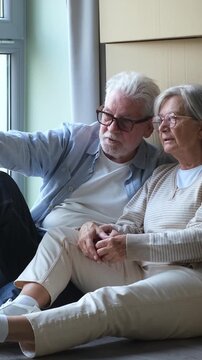 Loving senior couple sitting on the floor, relaxing and holding hands after moving into their new apartment, surrounded by cardboard boxes, talking and planning their future home together
