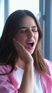 Frustrated young woman experiencing severe tooth pain, touching her cheek with a pained expression due to sensitive teeth, caries, or pulpitis problems while sitting indoors