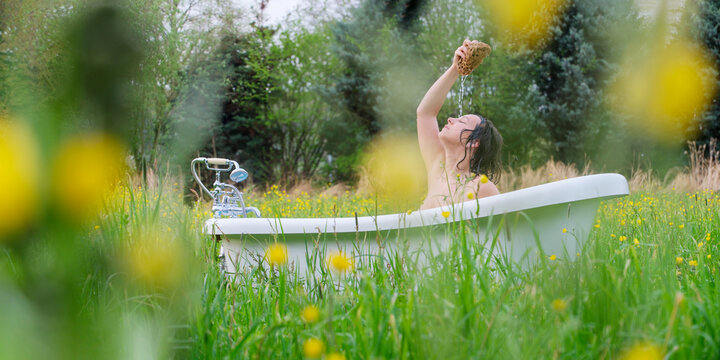 Woman bathing outdoors in a flowering meadow while pouring water from a natural sponge, creating a poetic atmosphere of freedom, freshness and serenity