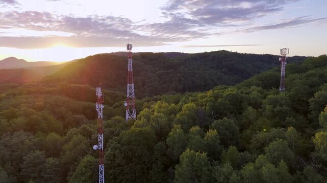 Aerial drone flight around 5G telecommunication towers above dense green forest at sunset with glowing sun rays and dramatic sky in nature landscape