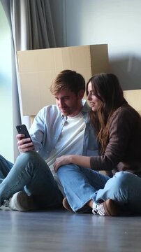 Happy young couple sitting on the floor of their new home, surrounded by unpacking boxes, while looking at a smartphone and planning their future together after moving in