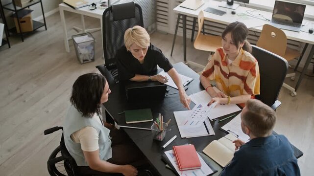 High angle view of young adult Caucasian woman in wheelchair joining diverse colleagues sitting at table while analyzing papers during office briefing