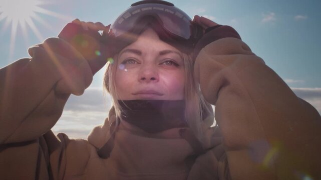 A beautiful girl puts on winter goggles or a mask for snowboarding. Big Blue eyes. Blond hair moving in the wind with slow motion. Getting ready for skiing on a snowy slope. Travel at the ski resort