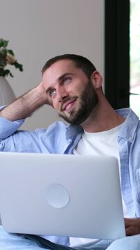 Relaxed young man with a beard smiling and daydreaming while working on his laptop, sitting comfortably on a couch in a modern living room, contemplating his future with satisfaction