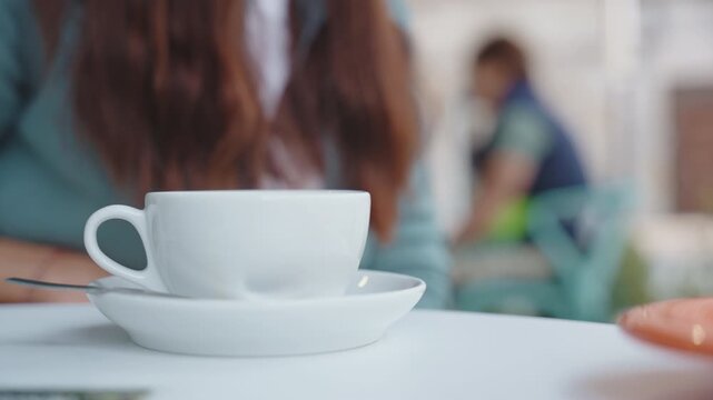 Close-up of a white coffee cup on a saucer resting on a table in a blurred cafe setting.