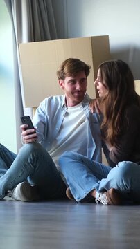 Happy young caucasian couple sitting on the floor of their new home, surrounded by cardboard boxes, looking at a smartphone to plan their future together and choose new furniture