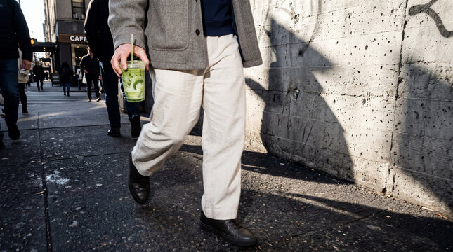 An elegant person in white linen trousers holding a green matcha latte while walking on a city street.