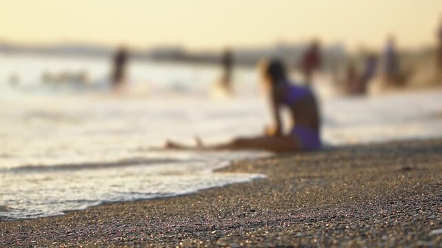 Warm golden hour light illuminates sparkling sand and gentle ocean waves as young person sits peacefully on shore, enjoying coastal atmosphere with blurred figures in distance. Low angle. Vacation hol
