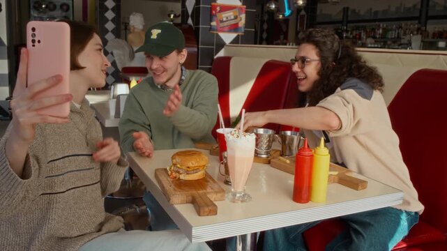 Medium shot of three Caucasian students eating out at diner, girl distracting friend while guy adding hot chili sauce to burger, then all laughing and filming reaction on smartphone
