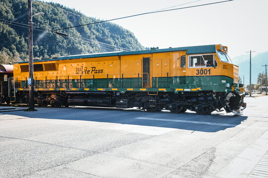 A yellow and green White Pass and Yukon Route diesel locomotive engine (Number 3001) crosses a street while a flagman directs traffic.