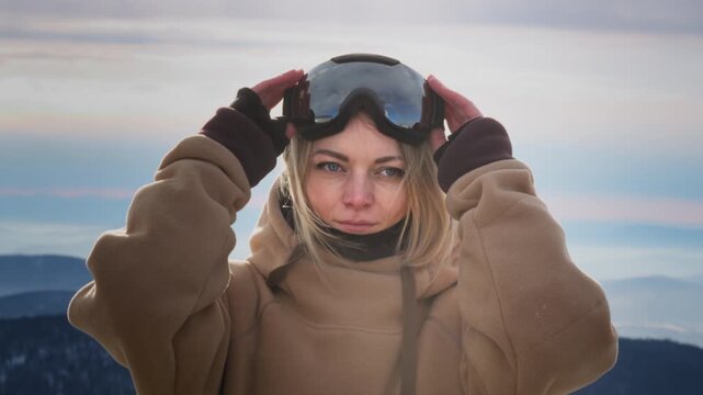 A beautiful girl puts on winter goggles or a mask for snowboarding. Big Blue eyes. Blond hair moving in the wind with slow motion. Getting ready for skiing on a snowy slope. Travel at the ski resort