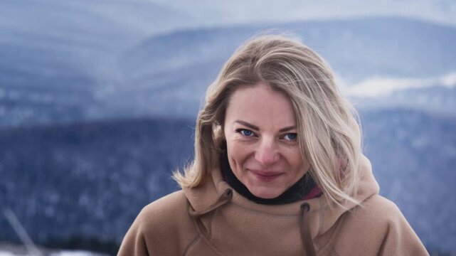 Portrait of a beautiful girl in a ski resort. Blond hair moving in the wind in slow mo. Puts on a hood of a hoodie. Mountain peaks in the snow. Winter travel at the ski resort. Blue big eyes