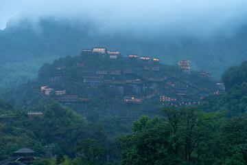 Fototapeta premium Traditional buildings with glowing lights stand on the green mountain slopes of Wangxian Valley Scenic Area in Jiangxi China during a foggy evening