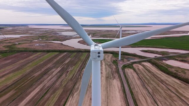 Cinematic drone shot aerial perspective of a field with windmills next to a river and the ocean in Nova Scotia. The landscape includes grass and water in the distance