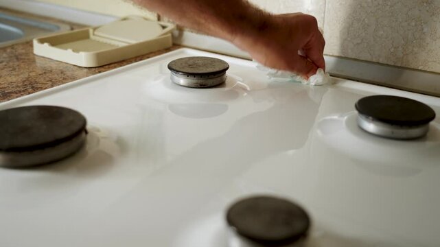 Man's hand cleans white gas stove top with a cloth in a bright kitchen