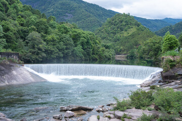 Fototapeta premium A scenic waterfall flows over a stone dam surrounded by lush green forests in the Wuyi Mountains of Fujian China