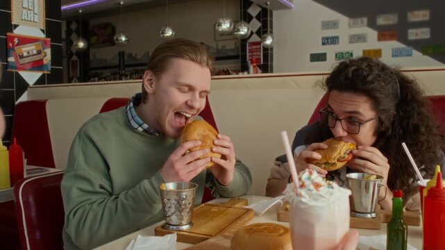 Medium shot of young Caucasian man enjoying tasty cheeseburger with fries, biting and chewing with closed eyes, while sitting in diner in company of friends, girl chatting and drinking milkshake