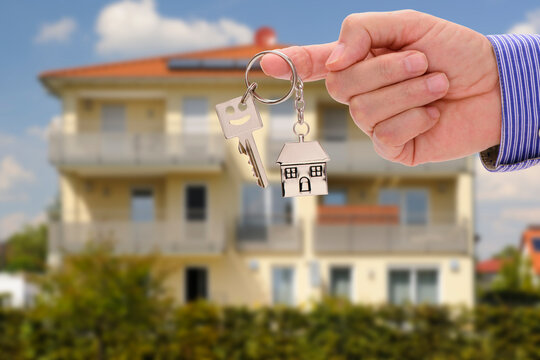 Eine Hand h&auml;lt einen Wohnungsschl&uuml;ssel vor einem neugebauten Haus als Symbol f&uuml;r ein Eigenheim - FOTOMONTAGE