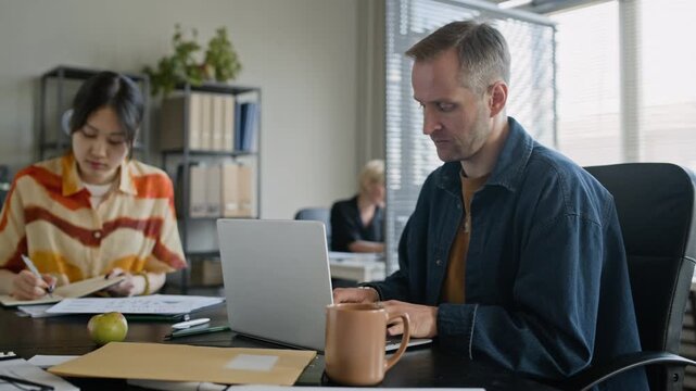 Medium handheld shot of biethnic coworkers sitting at table, doing report on laptop, reviewing documents and taking notes in modern office