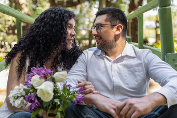 Fototapeta premium Happy couple sitting together in a garden holding a flower bouquet and sharing a warm romantic moment outdoors.