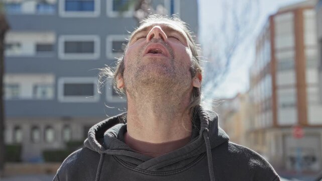 Man with exposed neck and stubble wearing a hoodie, looking upward in daylight on an urban street with apartment building behind; quiet contemplation.