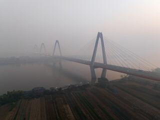 Naklejka premium Morning fog surrounds a long cable stayed bridge crossing a calm river above agricultural fields in northern Vietnam.
