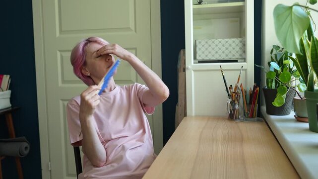 Tired middle aged woman sitting at table cooling herself with hand fan during heat indoor. Fatigue from high temperatures, heat exhaustion, coping with uncomfortable home climate. Lack air conditioner