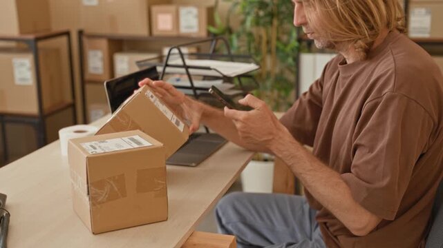 Young blond man scans parcel label with smartphone, hand on box and focused gaze while packing parcels in a building; focused productivity.