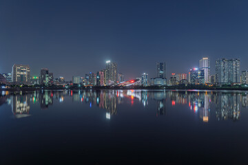 Fototapeta premium Illuminated bridge and modern skyline reflect across tranquil Cau Giay Lake during a peaceful Hanoi night scene.