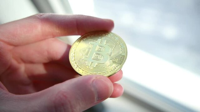 Human fingers holding shiny golden bitcoin coin against bright blurred background