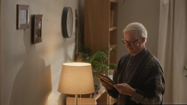 Side view medium shot of senior Caucasian man wearing glasses smiling while holding framed photograph in warm cozy apartment interior with soft lamp light
