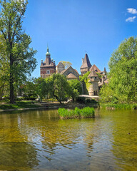 Obraz premium A daytime landscape view of the historic Vajdahunyad Castle located in the City Park. The architectural facade is reflected clearly on the calm water surface of the lake, surrounded by green summer tr