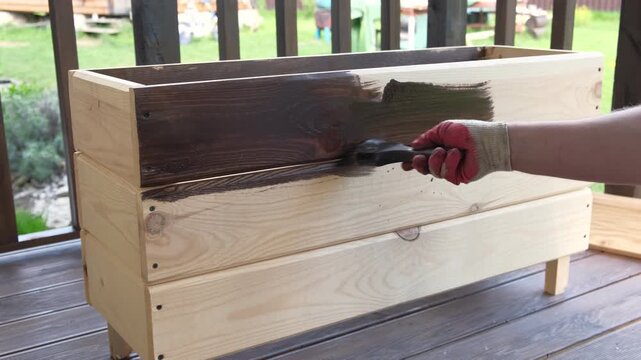 Brown oil for protection of wood by staining. Painting fence for protecting and pests. Woodworker applying finish to pine board box. Repairman varnishing with brush wooden fencing wall plank surface.