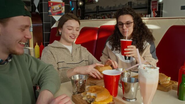 Medium shot of three young zoomers hanging out together at burger cafe, girl telling funny exam story and male friends rolling with laughter, then toasting each other with burgers, soda and milkshake
