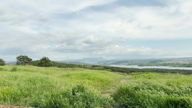 A vast green meadow of tall grass sways gently on a hill. The high field looks down upon a wide lake, dense forests, and faraway mountains under a bright, cloudy sky.