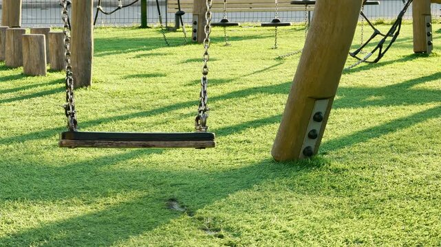 An empty wooden swing hangs from metal chains in a modern playground. It sits still over a vibrant green lawn of artificial turf. Long shadows stretch from the surrounding wooden play structures.