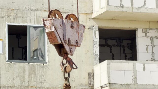 A large metal crane hook hangs near a raw apartment building wall. Active construction machinery runs continuously across the site. The heavy labor fills the area with a steady mechanical hum.