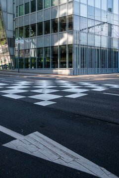 White road arrow cuts across a crosswalk beside a glass building, where urban street markings and facade reflections create architecture depth