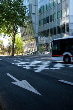 Road arrow points across an urban street toward a glass building, as crosswalk stripes, bus movement, and shadow define clean architecture