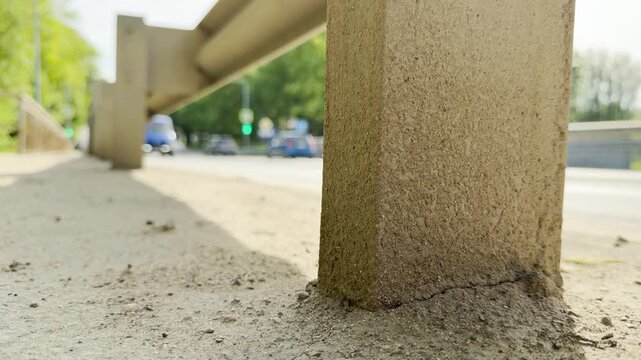 A white car drives quickly down a sunlit city street behind a solid concrete support pillar. Its swift forward motion produces a loud, continuous roar of traffic.